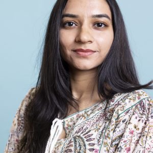Indian woman posing in a cute stylish outfit on camera and smiling Indian woman posing in a cute stylish outfit on camera and smiling, standing against blue background. Young lively person showing confidence during a photoshoot in the studio.
